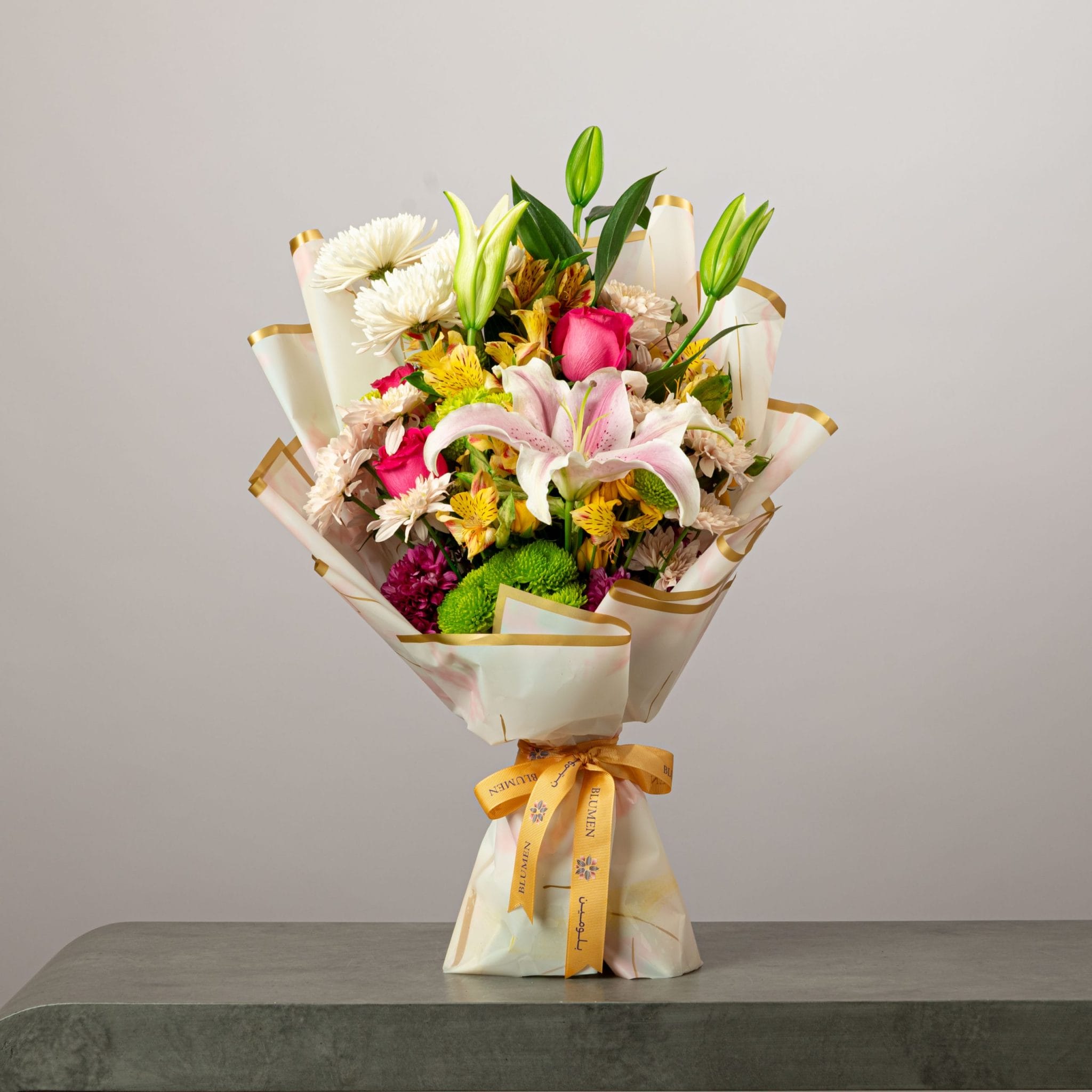Vibrant bouquet of pink lilies, roses, and mixed flowers wrapped in white and gold paper with a yellow ribbon.