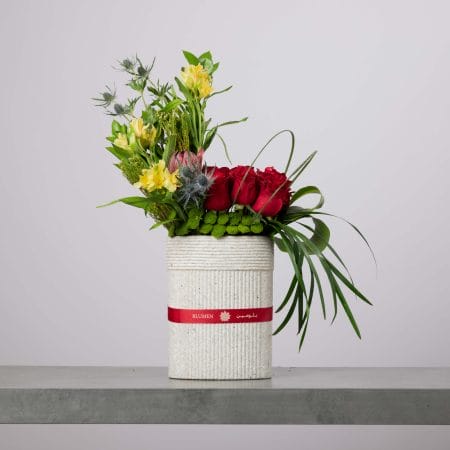 Tall textured stone-like cylinder vase with a red Blumen ribbon, arranged with red roses, yellow blooms, blue thistle accents, green button flowers, and cascading greenery against a light grey background.
