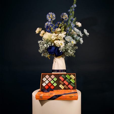 Elegant floral arrangement featuring deep blue, ivory, and soft blue blossoms with lush greenery, arranged in a sculptural ceramic vase.