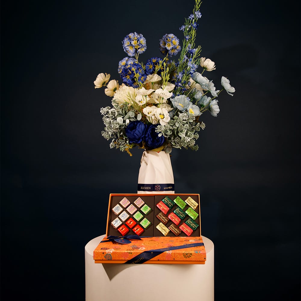 Elegant floral arrangement featuring deep blue, ivory, and soft blue blossoms with lush greenery, arranged in a sculptural ceramic vase.