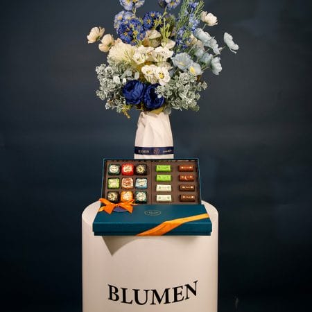 Elegant floral arrangement featuring deep blue, ivory, and soft blue blossoms with lush greenery, arranged in a sculptural ceramic vase.