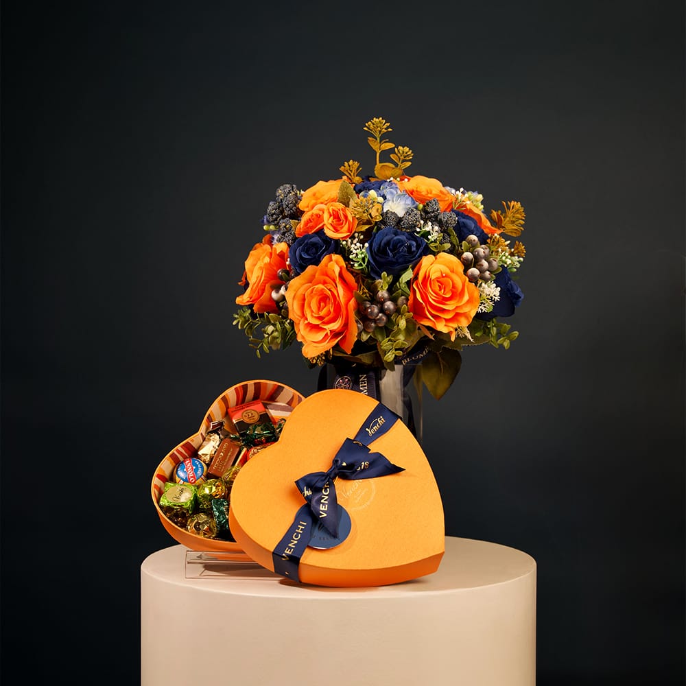 Floral arrangement with bright amber-orange roses, deep blue flowers, and greenery arranged in a clear glass vase with decorative ribbon.