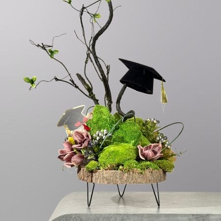 A modern botanical arrangement featuring preserved green moss, sculptural branches, floral accents, and graduation-themed decorations on a wooden display stand.