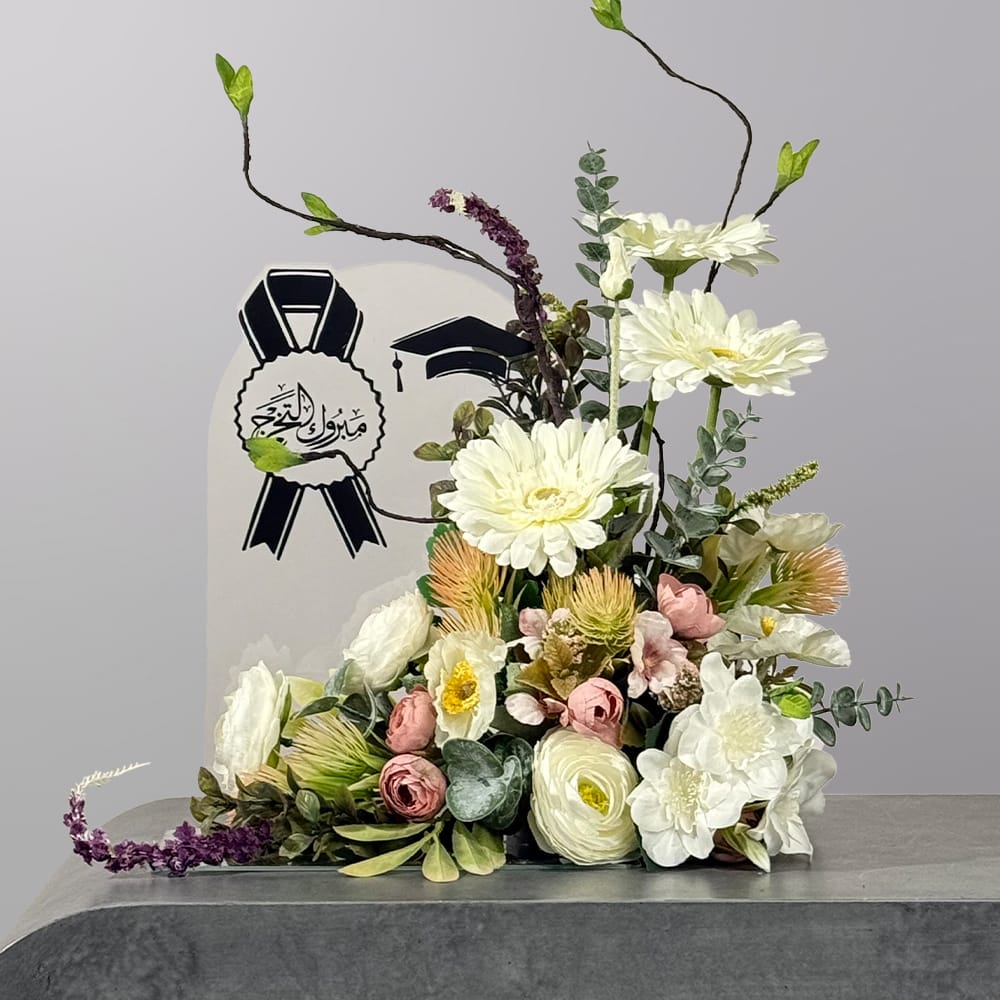 A sophisticated graduation-themed floral arrangement featuring white blooms, blush roses, eucalyptus, and artistic branches beside a congratulatory graduation sign.