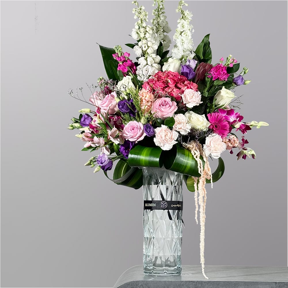 A luxurious floral arrangement featuring pink roses, white blooms, purple flowers, and vibrant greenery arranged in a tall crystal-style glass vase.