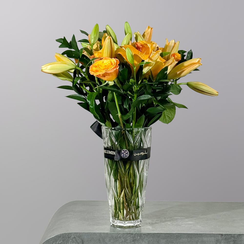 A refined floral arrangement featuring golden yellow roses and soft yellow lilies arranged in a crystal glass vase with rich green foliage.