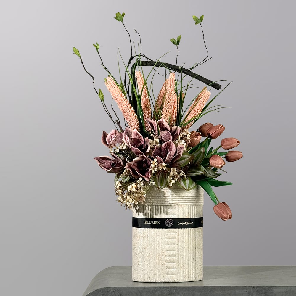 A modern floral arrangement featuring mocha-toned tulips, muted orchids, textured pink blooms, and artistic branches in a tall ceramic vase.