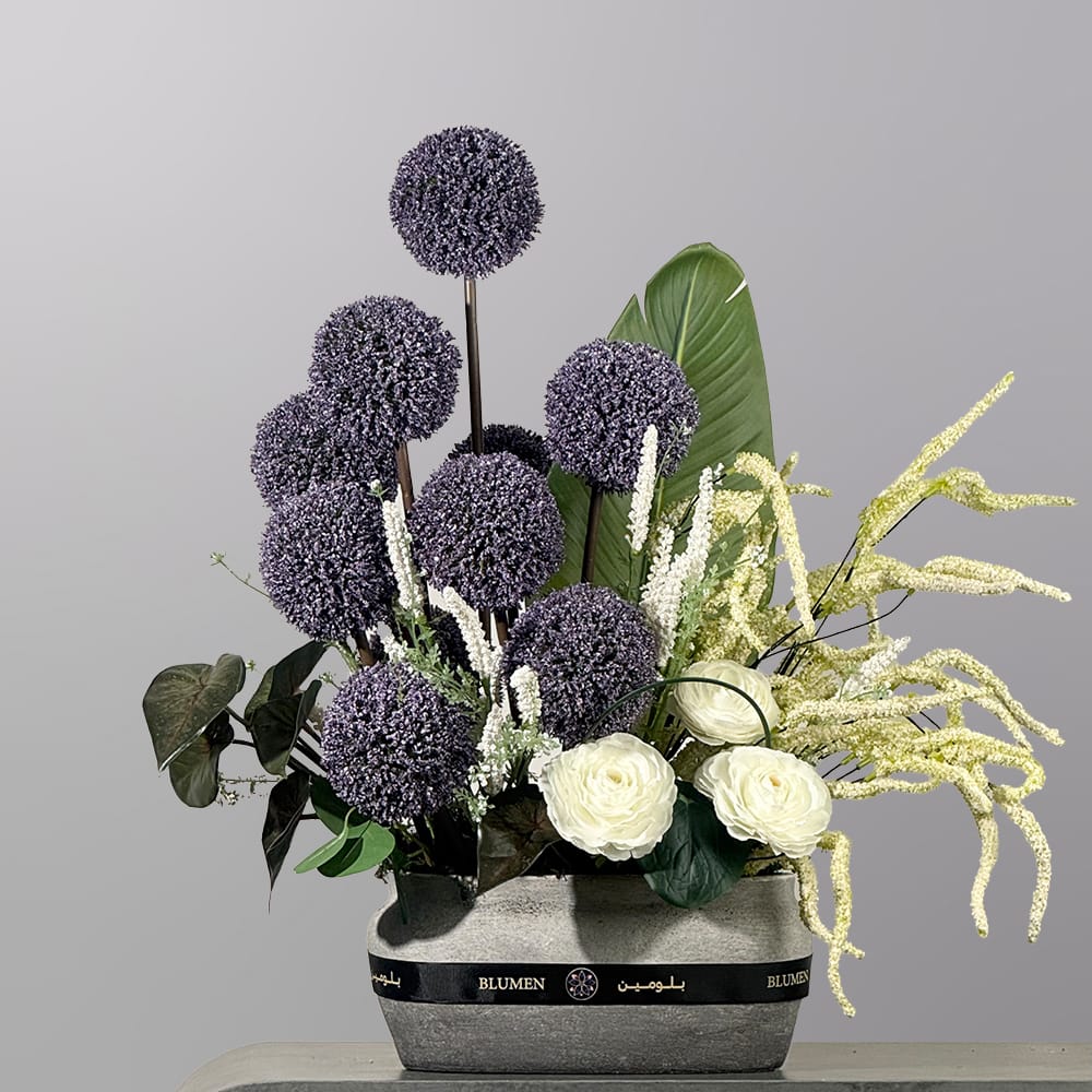 A contemporary floral arrangement featuring spherical purple allium flowers, white roses, textured ivory blooms, and tropical greenery in a modern stone-textured vase.