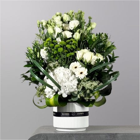 A luxurious white floral arrangement featuring hydrangeas, roses, lisianthus, green button chrysanthemums, and white gerberas arranged in a modern white vase with flowing greenery accents.