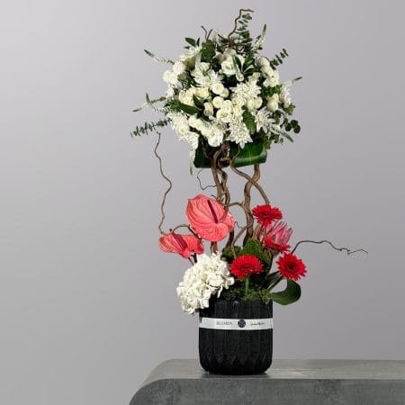 A luxurious tall floral arrangement featuring white chrysanthemums, white roses, pink anthuriums, red gerberas, hydrangeas, twisting branches, and moss details in a modern black vase.