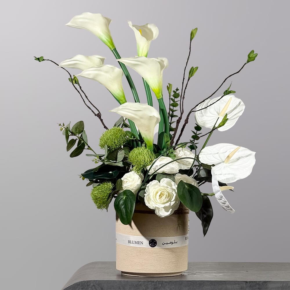 An elegant floral arrangement featuring white calla lilies, white anthuriums, white roses, green chrysanthemums, and artistic branches arranged in a soft beige vase.