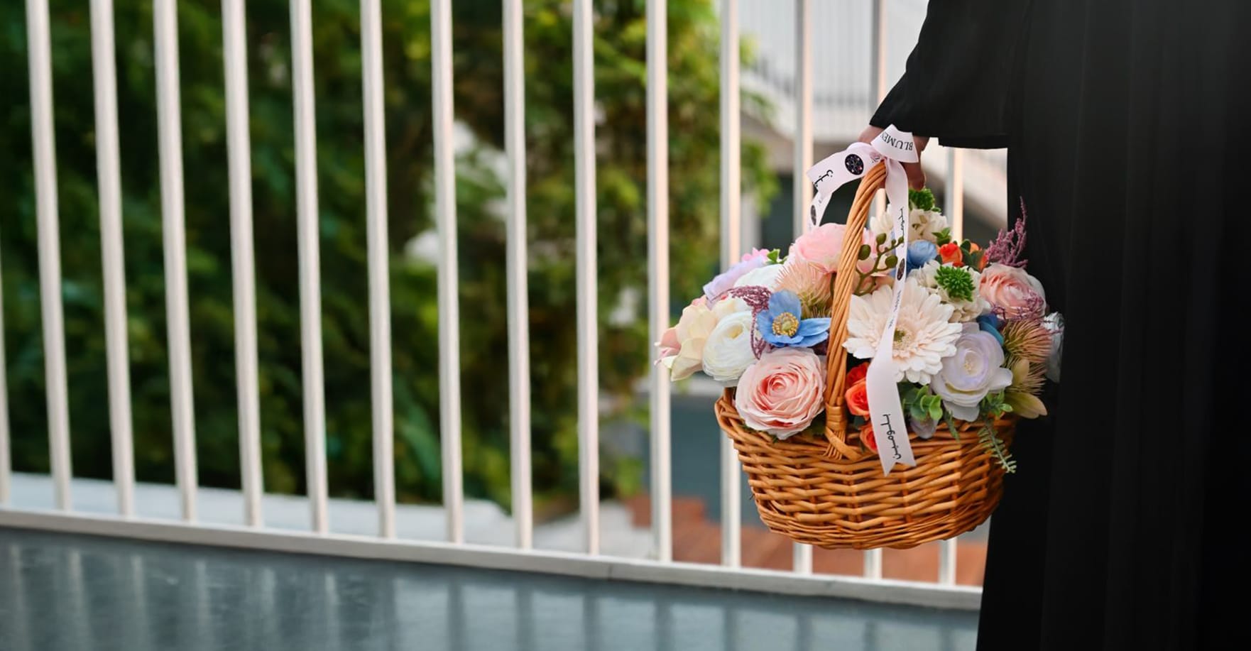 Woman wearing a black abaya holding a woven basket with a bouquet of pink roses and pastel flowers on an outdoor balcony