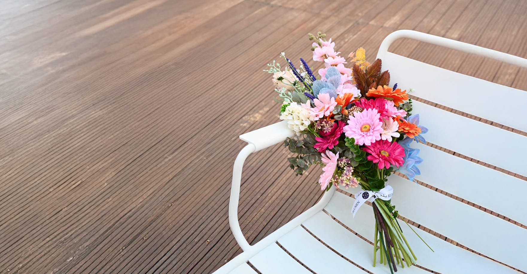 Vibrant bouquet of pink and orange gerberas with blue accents placed on an outdoor white bench
