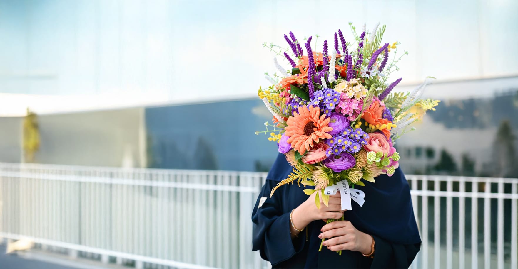 Woman wearing a navy abaya holding a vibrant bouquet of purple veronicas, orange gerberas and pink roses