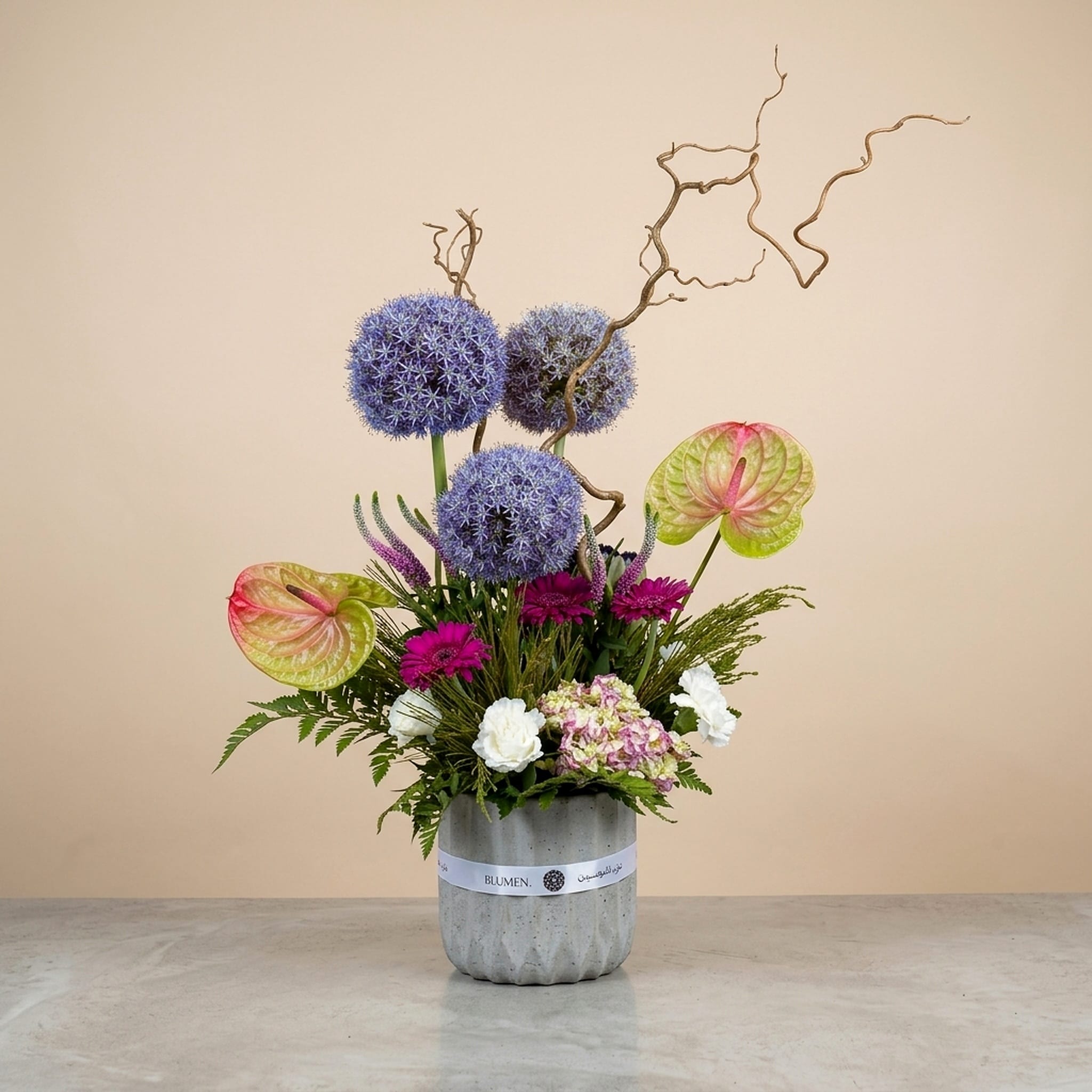 The Starlit Mirage floral arrangement featuring blue allium blooms, pastel anthuriums, pink hydrangea, magenta gerberas, and artistic curly branches in a modern stone vase.