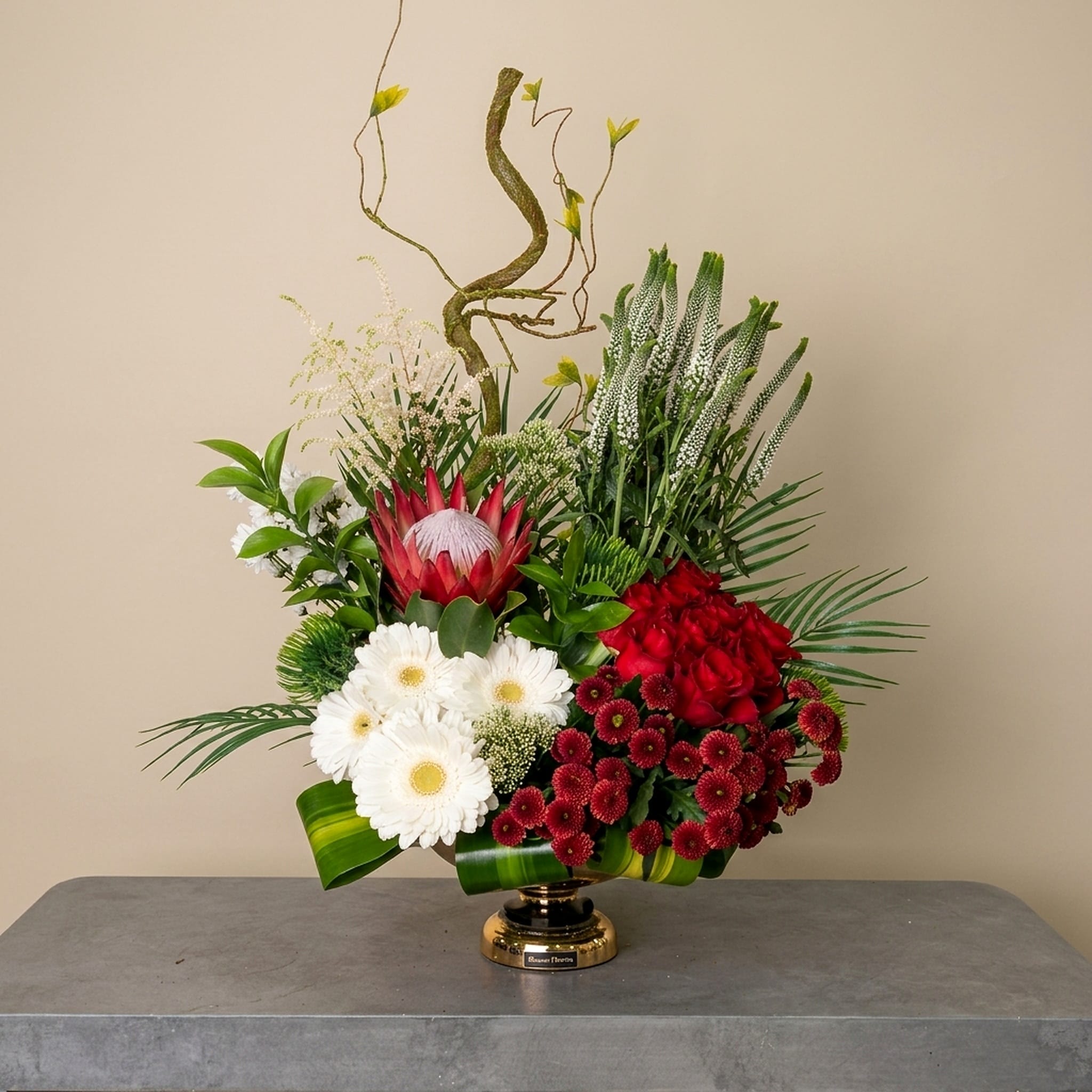 An artistic floral arrangement featuring a king protea, red roses, burgundy chrysanthemums, white gerberas, and architectural greenery displayed in a decorative pedestal bowl.
