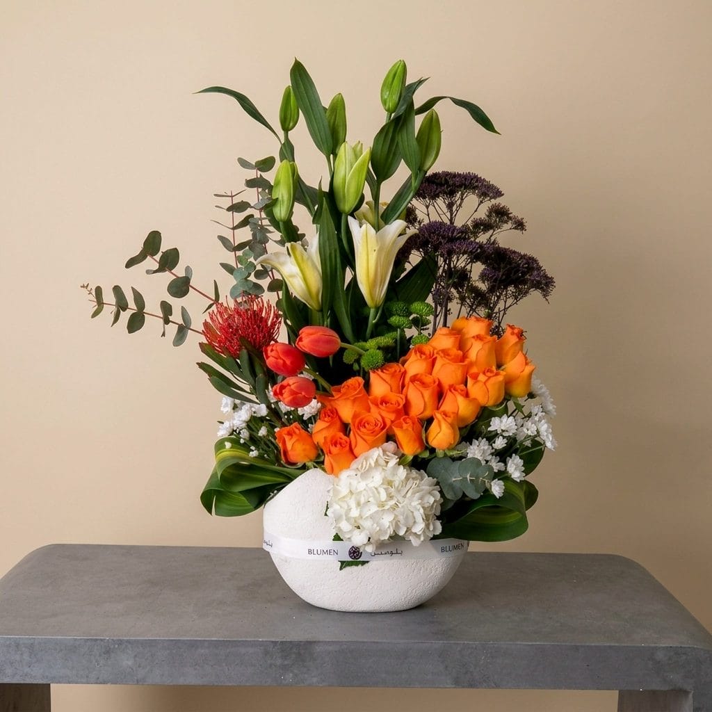 A luxurious floral arrangement featuring orange roses, white lilies, tulips, hydrangeas, and exotic foliage arranged in a contemporary white ceramic bowl.