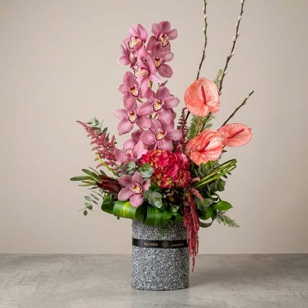 A luxury floral arrangement featuring pink cymbidium orchids, blush anthuriums, hydrangeas, and cascading botanical accents arranged in a contemporary textured vase.