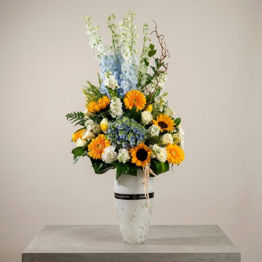 A luxury floral arrangement featuring sunflowers, yellow gerberas, blue hydrangeas, white roses, and delphiniums arranged in a tall white vase.