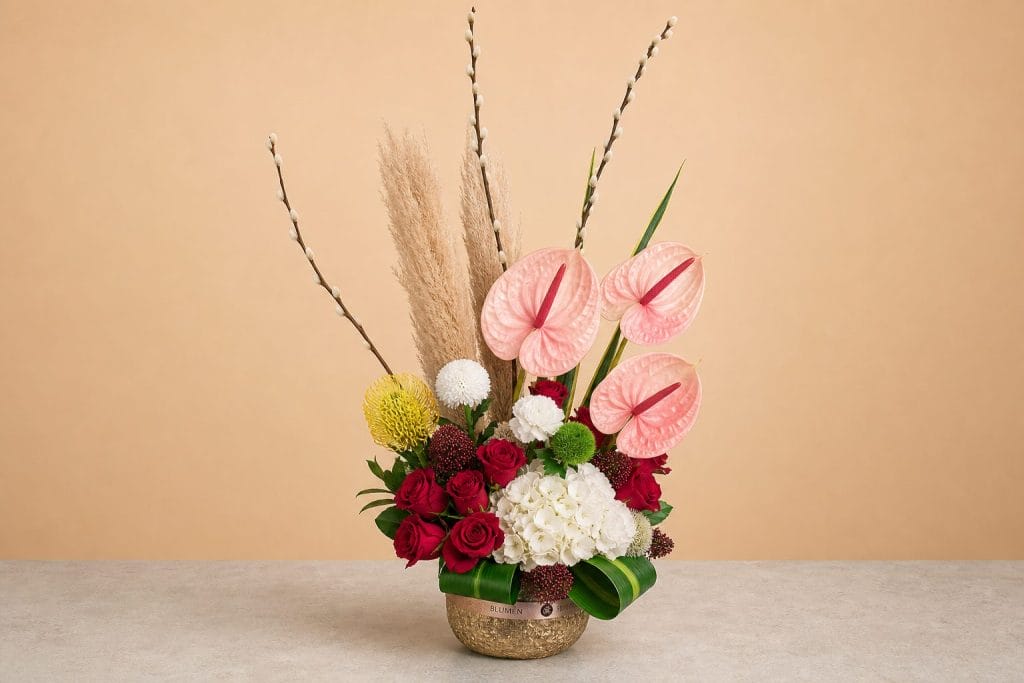 Luxury floral arrangement featuring blush pink anthuriums, red roses, white hydrangea, pampas grass, pussy willow branches, and seasonal blooms in a textured golden vase.
