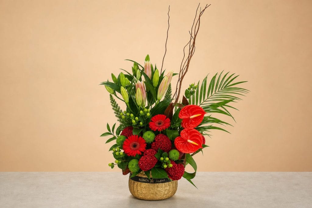 Luxury floral arrangement featuring red anthuriums, red gerbera daisies, red cockscomb blooms, pink lilies, green button chrysanthemums, tropical foliage, and decorative branches in a gold vase.