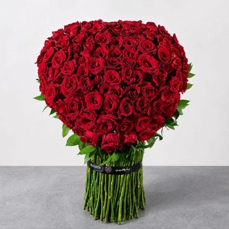 The Crimson Devotion heart-shaped arrangement featuring premium red roses with lush green foliage and exposed stems.