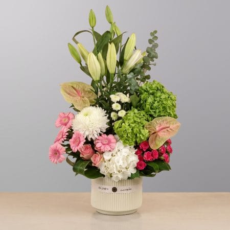A sophisticated floral arrangement featuring premium blooms, delicate seasonal flowers, lush greenery, and botanical accents, elegantly arranged in a decorative container.