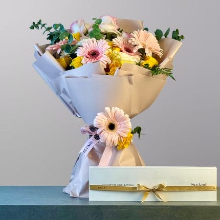 A soft and elegant bouquet of peach gerberas, yellow roses, pink and cream roses, and delicate greenery wrapped in luxurious cream paper with a white box of chocolates.