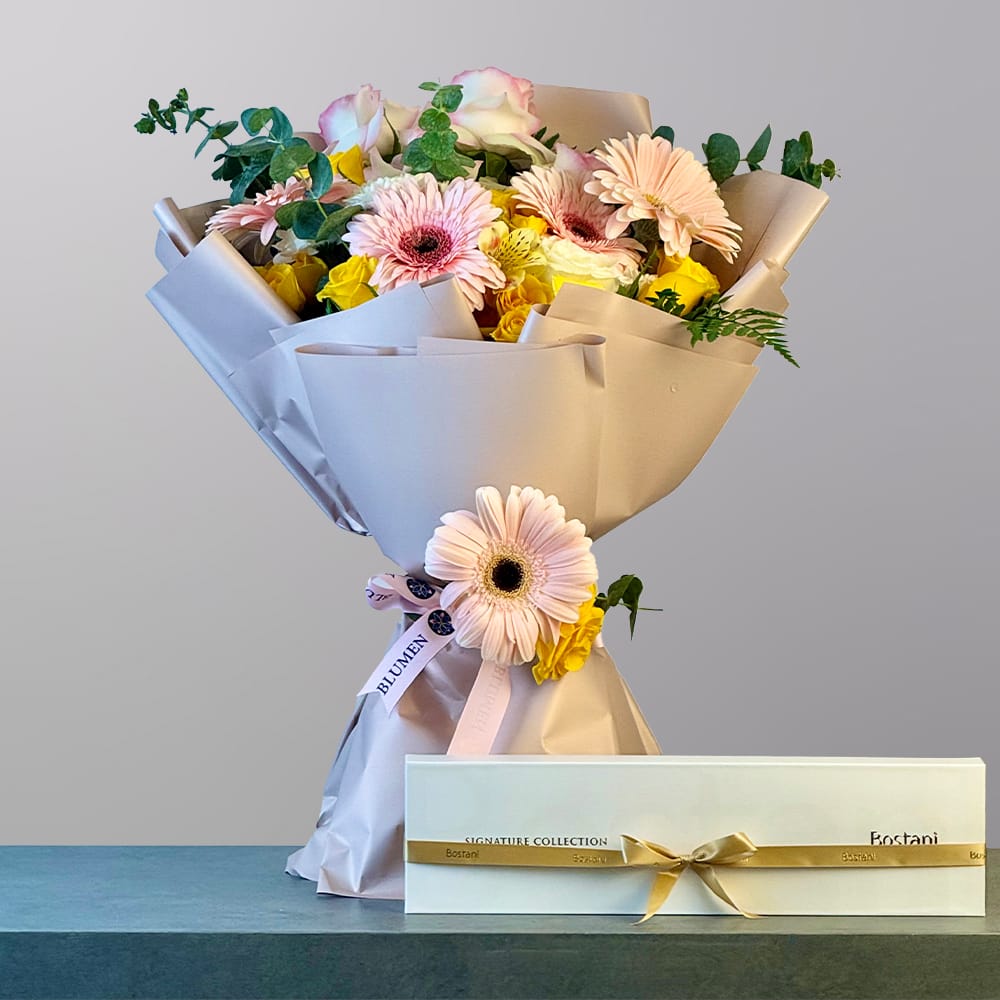 A soft and elegant bouquet of peach gerberas, yellow roses, pink and cream roses, and delicate greenery wrapped in luxurious cream paper with a white box of chocolates.