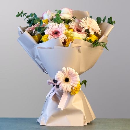 A soft and elegant bouquet of peach gerberas, yellow roses, pink and cream roses, and delicate greenery wrapped in luxurious cream paper.