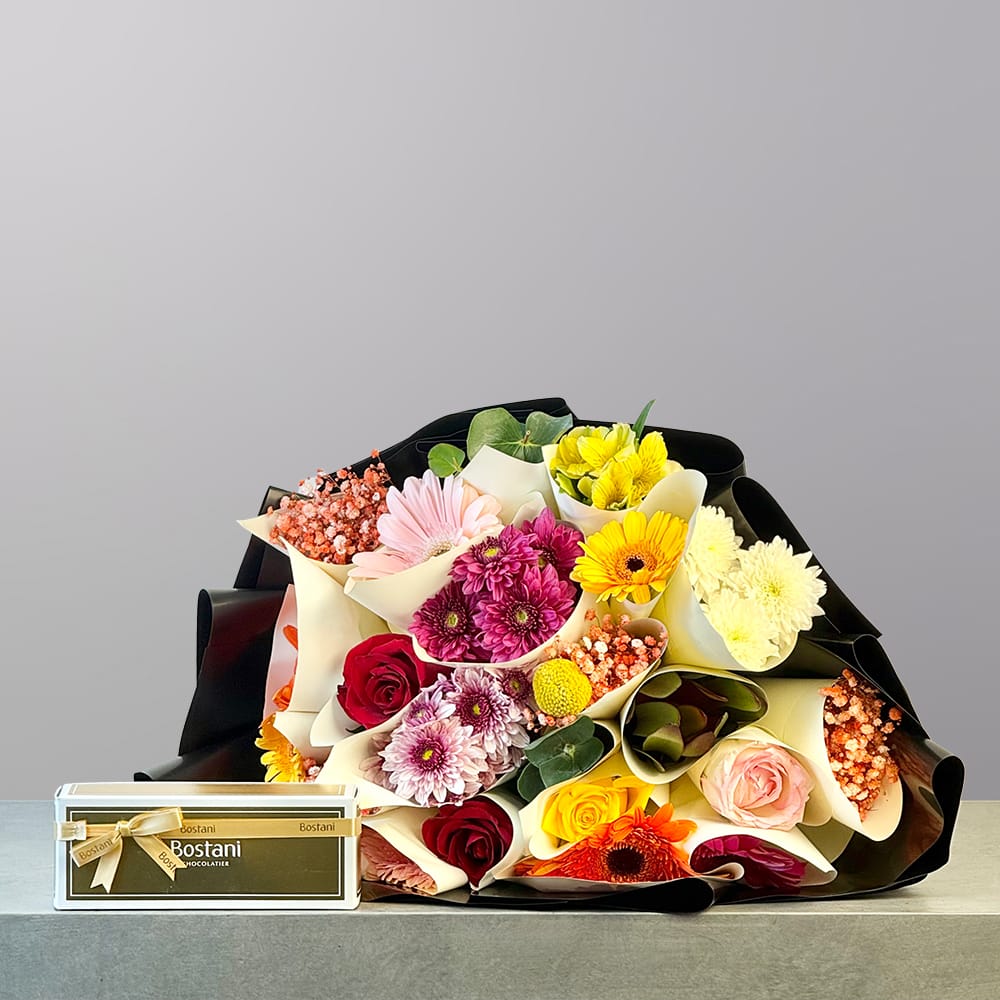 A vibrant hand-tied bouquet featuring pink, yellow, orange, red, white, and peach flowers wrapped in elegant black and cream paper.