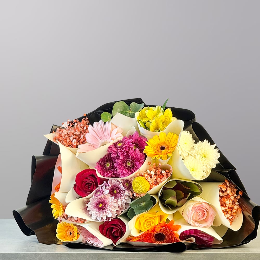 A vibrant hand-tied bouquet featuring pink, yellow, orange, red, white, and peach flowers wrapped in elegant black and cream paper.