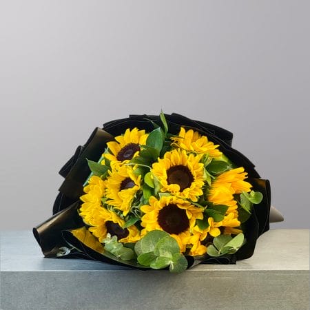 A lush bouquet of bright yellow sunflowers and fresh green foliage wrapped in elegant black paper.