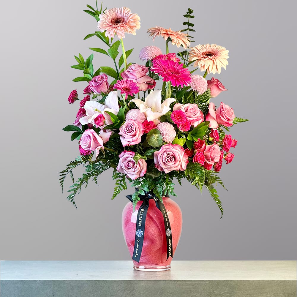 A lush bouquet of pink and peach gerbera daisies, roses, lilies, and chrysanthemums arranged in a pink glass vase with green foliage.
