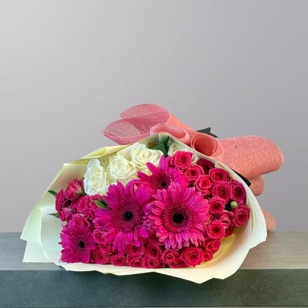 A vibrant bouquet of fuchsia gerbera daisies, hot pink spray roses, and creamy white roses wrapped in elegant coral-pink paper.