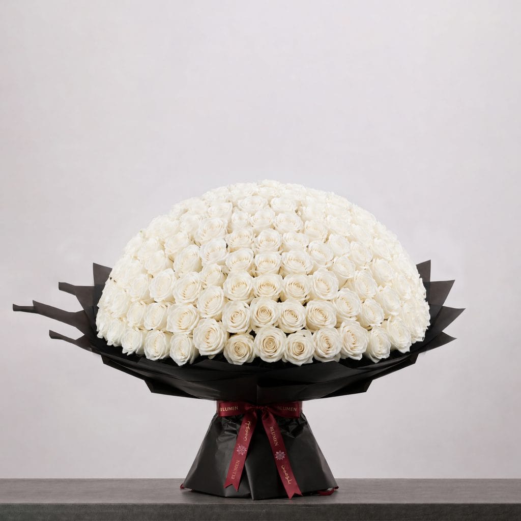 A large bouquet of elegant white roses arranged in a full, rounded shape, wrapped in sophisticated black paper and finished with a refined ribbon.