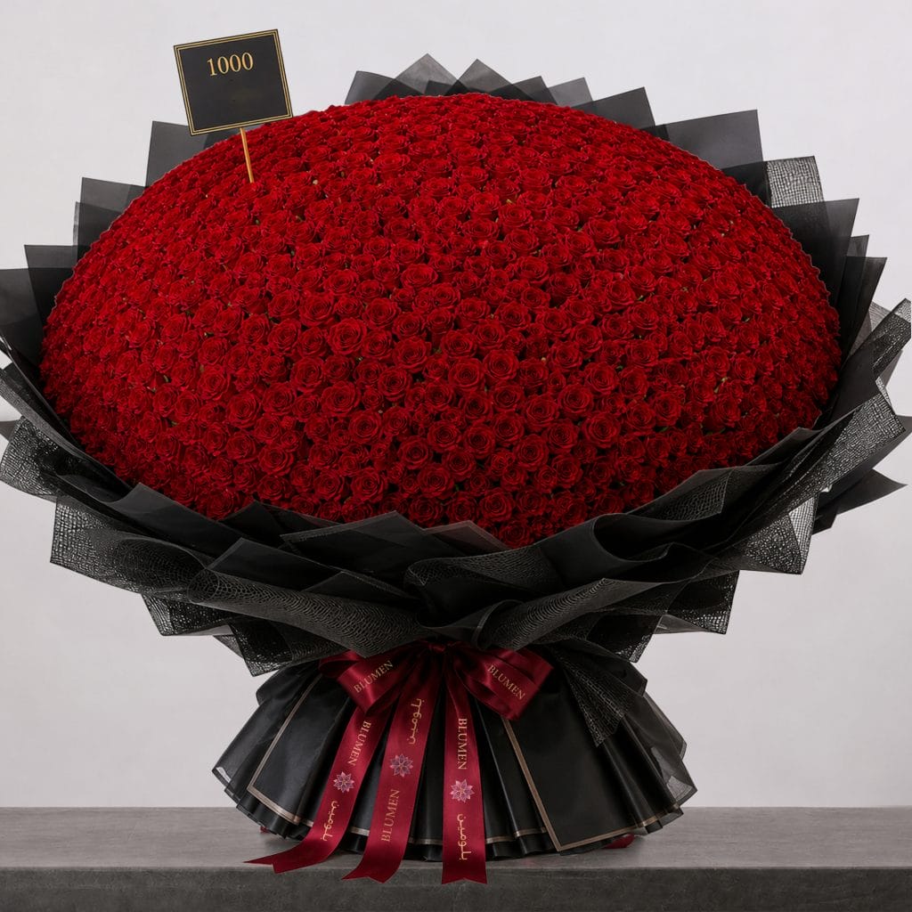 A large bouquet of deep red roses arranged in a full, rounded shape, wrapped in elegant black paper and finished with a burgundy ribbon.