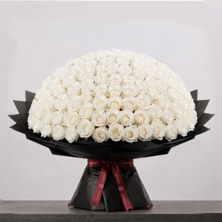 A large bouquet of elegant white roses arranged in a full, rounded shape, wrapped in sophisticated black paper and finished with a refined ribbon.