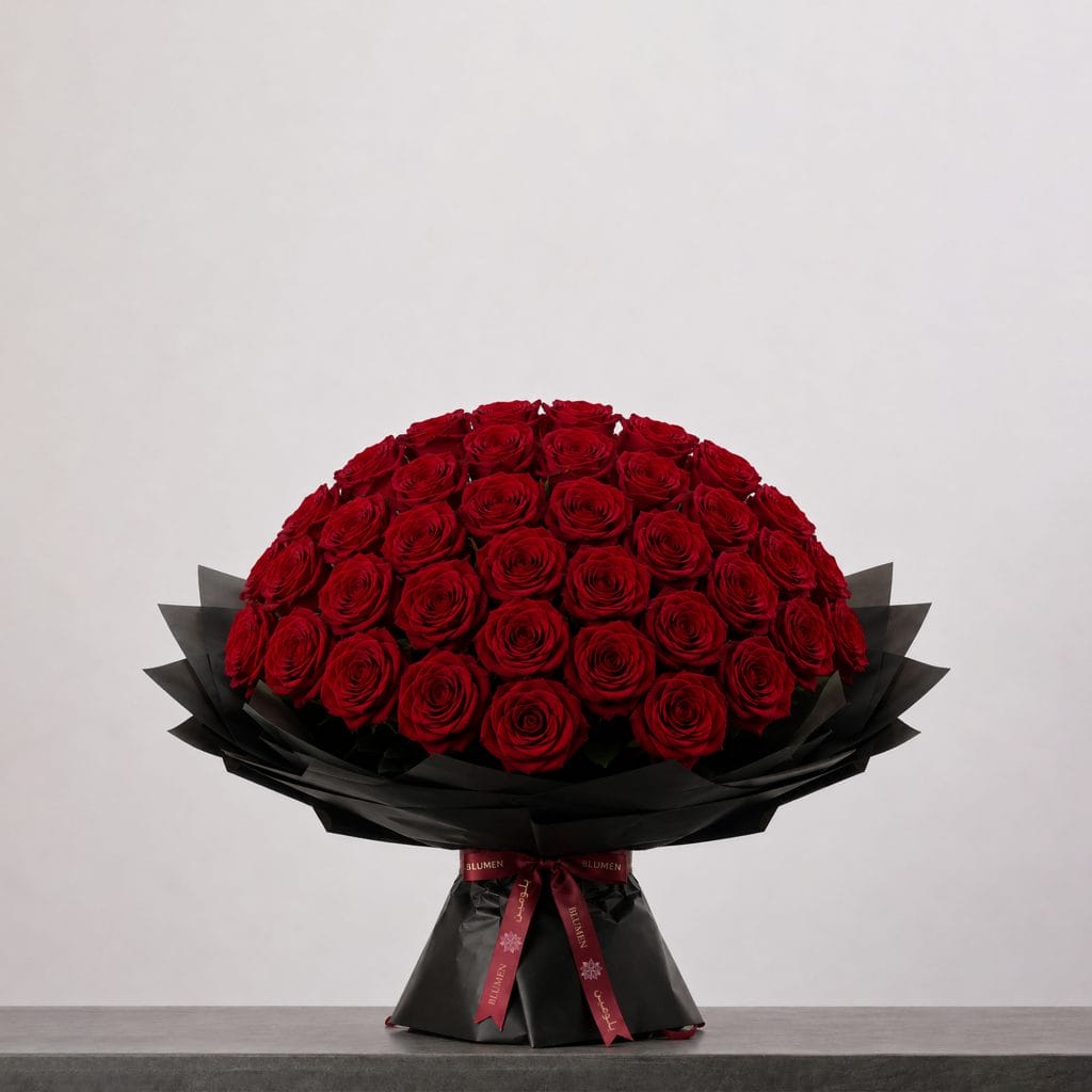A large bouquet of deep red roses arranged in a full, rounded shape, wrapped in elegant black paper and finished with a burgundy ribbon.