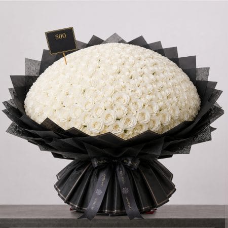 A large bouquet of elegant white roses arranged in a full, rounded shape, wrapped in sophisticated black paper and finished with a refined ribbon.